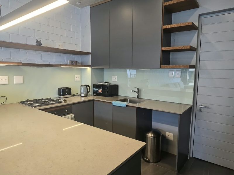 Modern kitchen featuring dark grey cabinetry, stone countertops, gas hob, under-counter oven, and stylish floating shelves with integrated lighting.