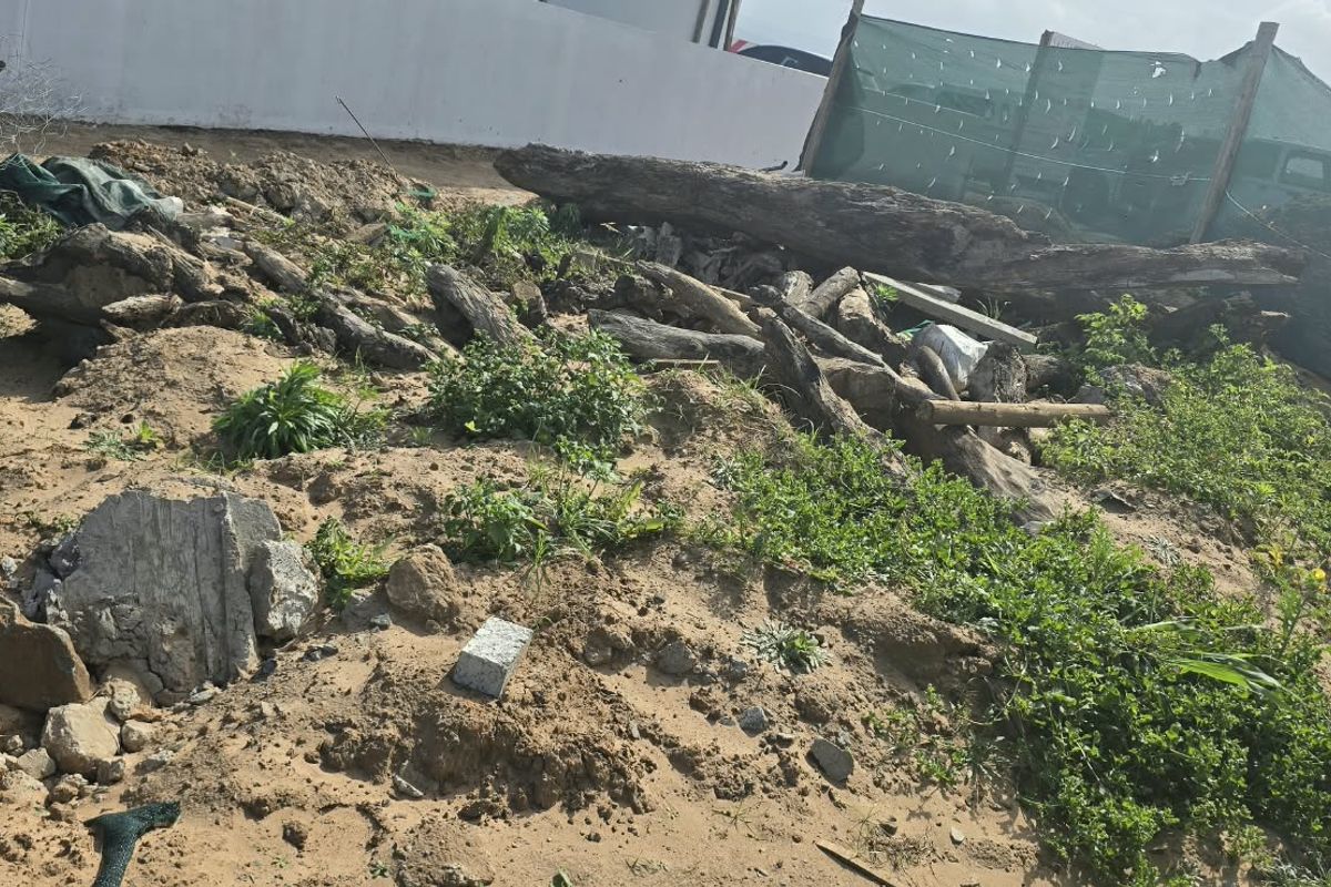 A view of a vacant residential plot featuring sandy terrain, scattered debris, and temporary boundary fencing with shade netting.