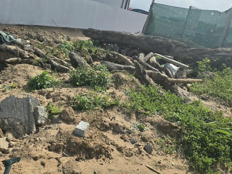 A view of a vacant residential plot featuring sandy terrain, scattered debris, and temporary boundary fencing with shade netting.