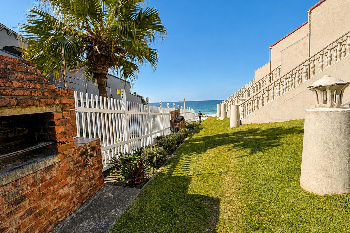 Rear garden area featuring a brick built-in braai, manicured lawn, and white picket fencing, with direct access to the beach.