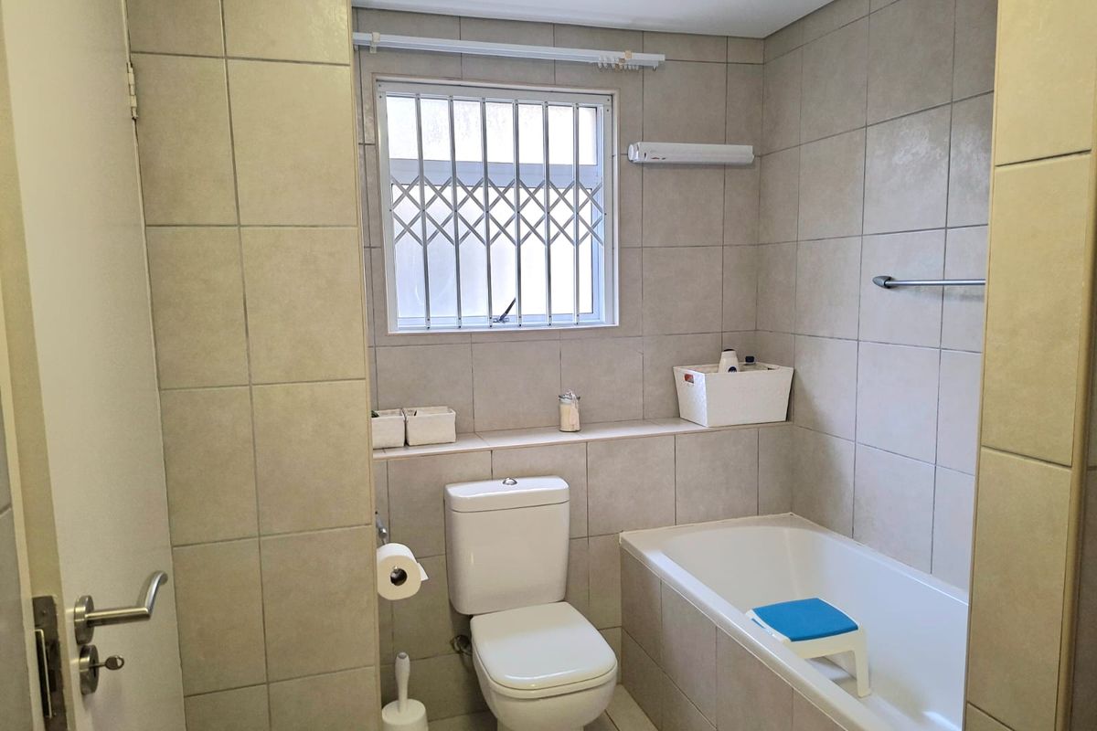A functional bathroom featuring neutral wall tiles, a built-in bathtub, a standard toilet, and a window fitted with a security gate.