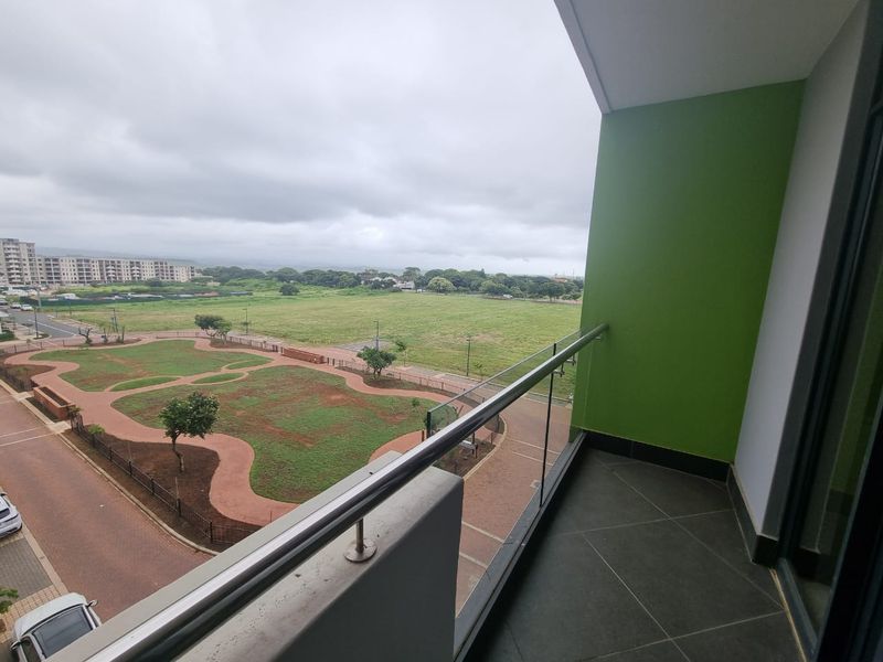 A modern apartment balcony featuring a glass balustrade, stainless steel handrail, and tiled flooring, offering an elevated view over the surrounding estate and parkland.