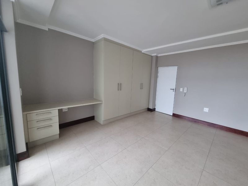 A bright, empty bedroom featuring neutral tiled flooring, built-in cupboards, and a convenient integrated desk area.