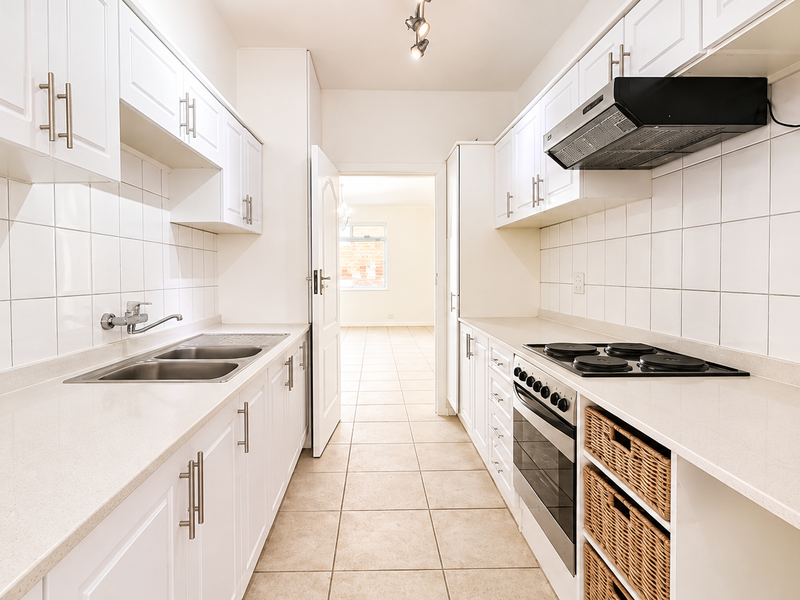 A clean, galley-style kitchen featuring white cabinetry, stone countertops, integrated oven and hob, and tiled finishes throughout.