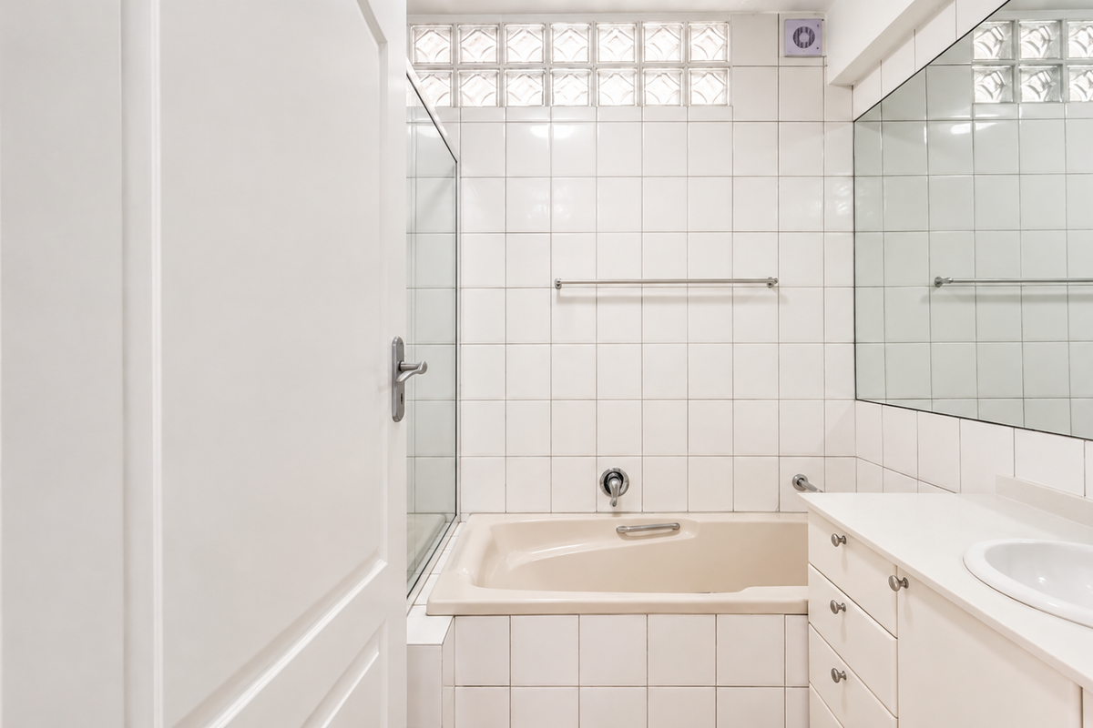A functional bathroom featuring a built-in bathtub, vanity cabinet with integrated sink, glass shower screen, and a glass block window for natural light.