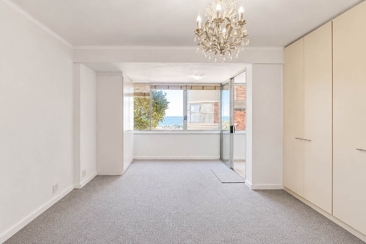 A bright, empty bedroom featuring neutral carpeting, extensive built-in wardrobe storage, and a sliding glass door leading to an outdoor balcony with ocean views.