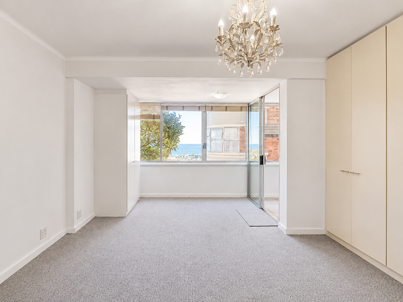 A bright, empty bedroom featuring neutral carpeting, extensive built-in wardrobe storage, and a sliding glass door leading to an outdoor balcony with ocean views.