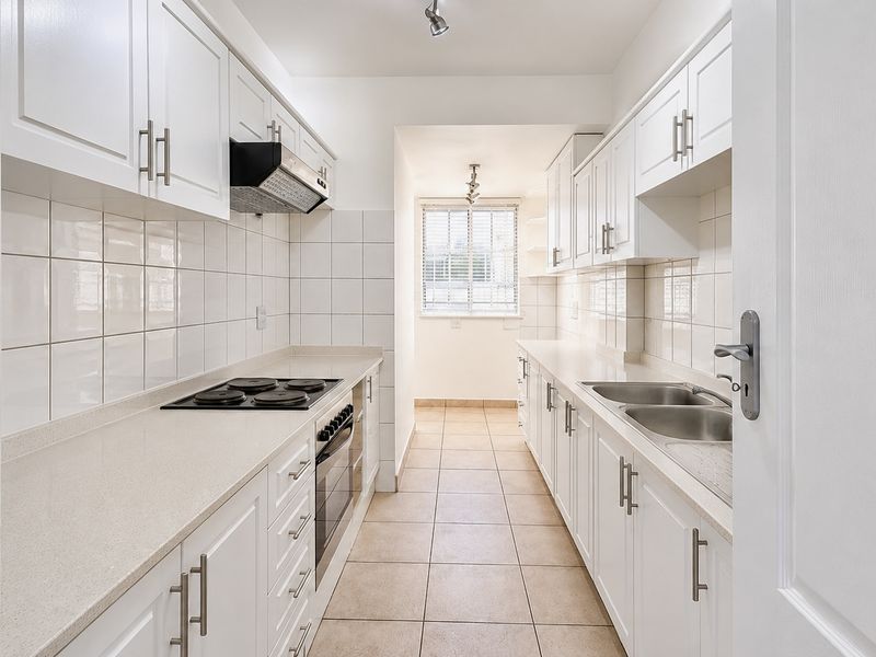 A clean, modern galley-style kitchen featuring white cabinetry, stone countertops, integrated appliances, and neutral tiled flooring.