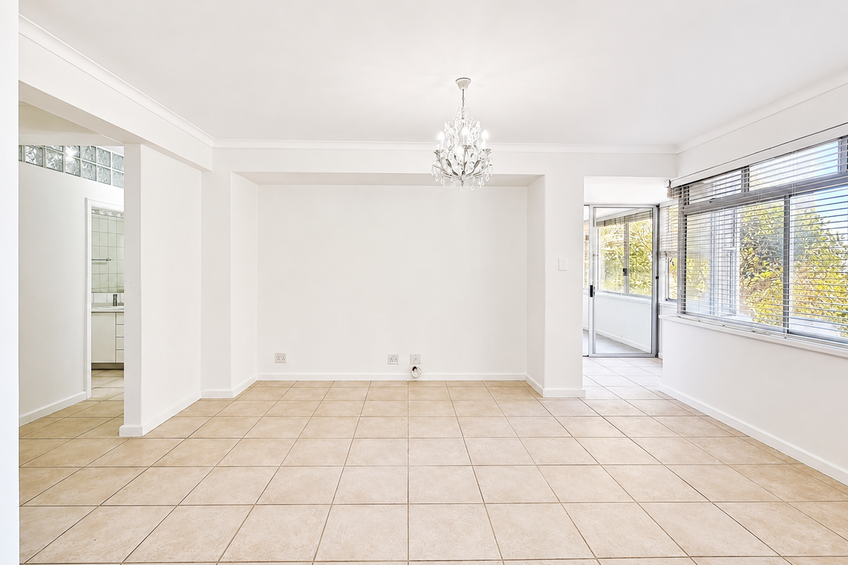 A bright, open-plan living area featuring neutral ceramic floor tiles, a decorative chandelier, and large windows with venetian blinds, offering a clean, versatile space.
