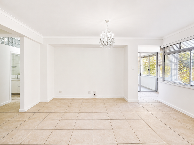 A bright, open-plan living area featuring neutral ceramic floor tiles, a decorative chandelier, and large windows with venetian blinds, offering a clean, versatile space.