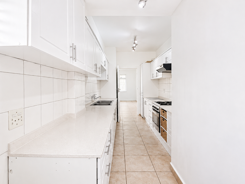 A clean, galley-style kitchen featuring white cabinetry, stone countertops, built-in oven, hob, and tiled finishes throughout.
