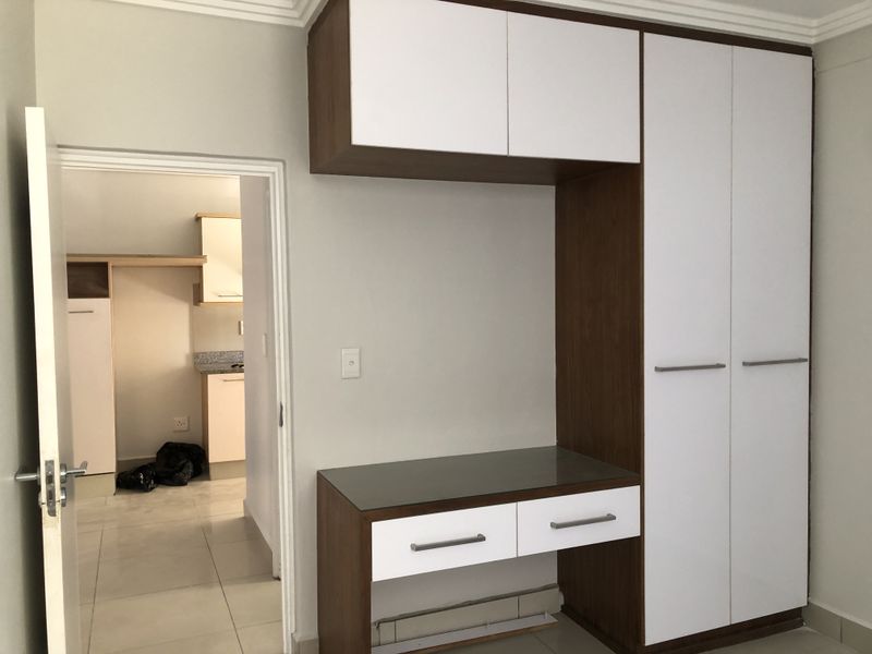 A clean bedroom featuring a modern built-in wardrobe and integrated vanity unit, with tiled flooring and a view into the adjacent kitchen area.
