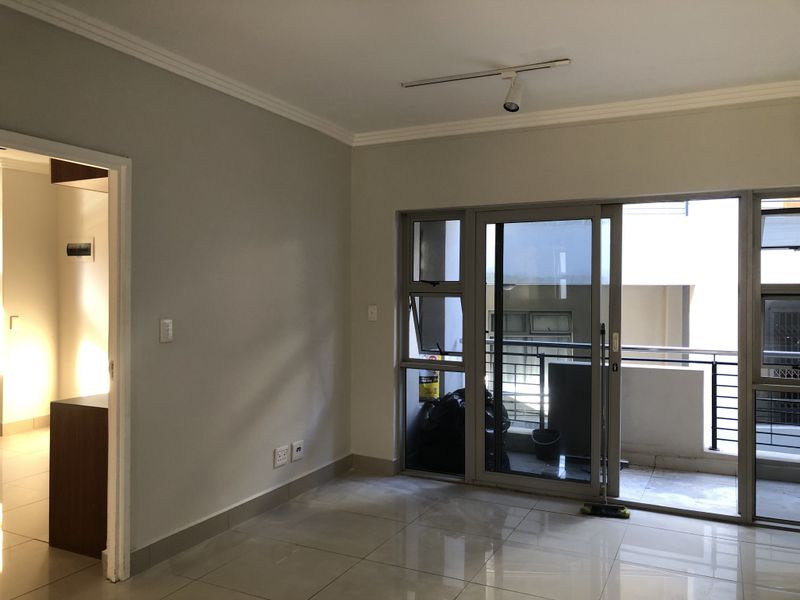 A bright, empty living area featuring neutral tiled flooring, track lighting, and a sliding glass door leading to an outdoor balcony.