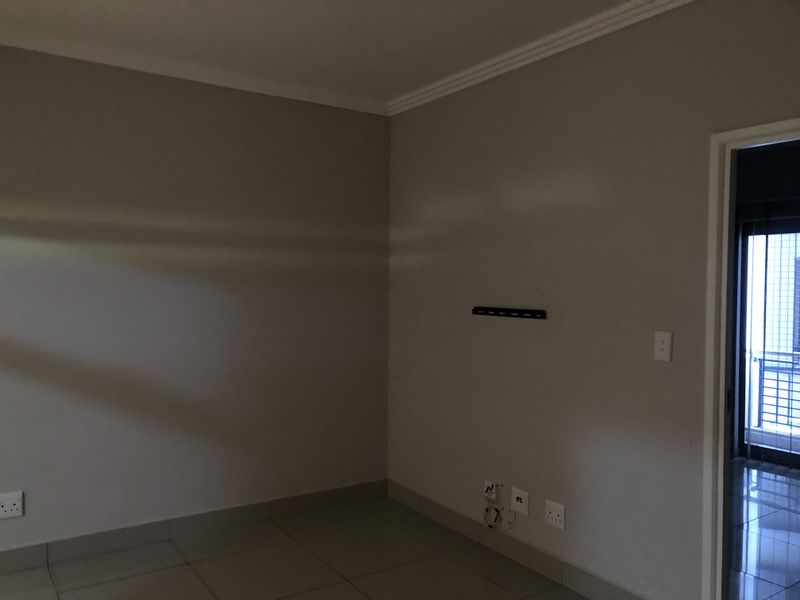 An empty living room area featuring tiled flooring, neutral wall paint, coving, and integrated wall-mounted TV bracket with electrical and data connectivity.