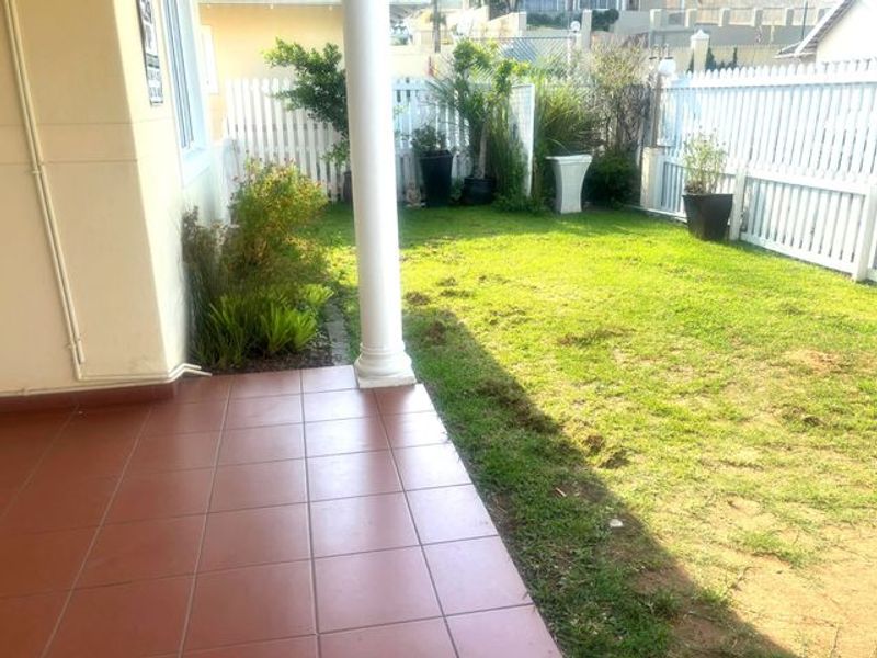 A rear garden area featuring a tiled patio, a white picket fence, and a lawn with some patchy areas.