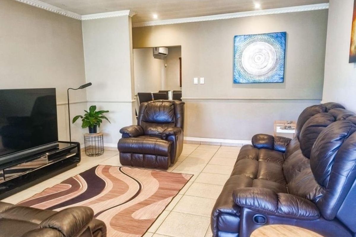 A comfortable living room featuring neutral wall tones, tiled flooring, crown molding, and recessed lighting, with an adjacent dining area visible in the background.
