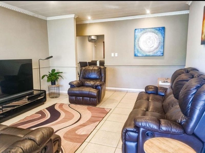 A comfortable living room featuring neutral wall tones, tiled flooring, crown molding, and recessed lighting, with an adjacent dining area visible in the background.