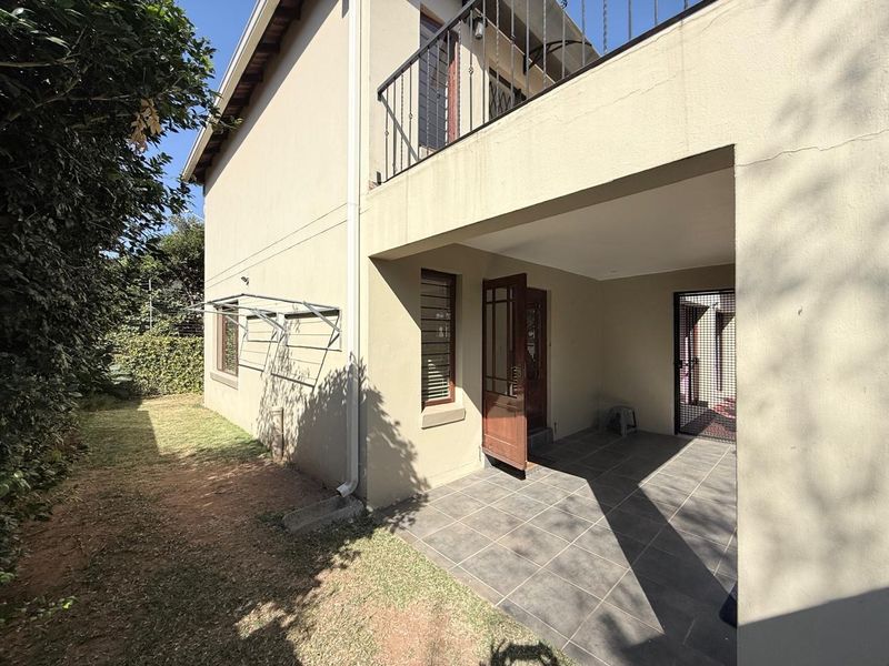 A rear exterior view showing a covered tiled patio, a small garden area, and an upper-level balcony with metal railings.
