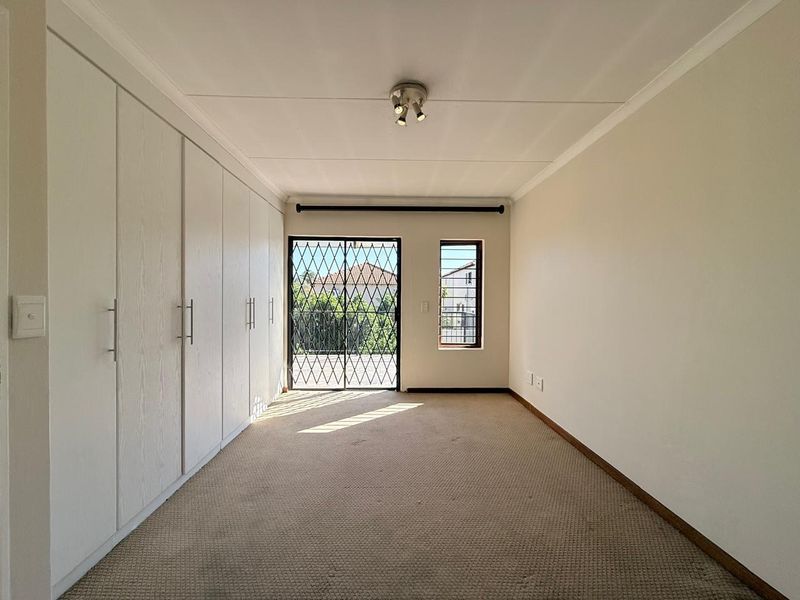 A spacious, empty bedroom featuring extensive built-in cupboards, carpeted flooring, and a sliding door with a security gate leading to the exterior.