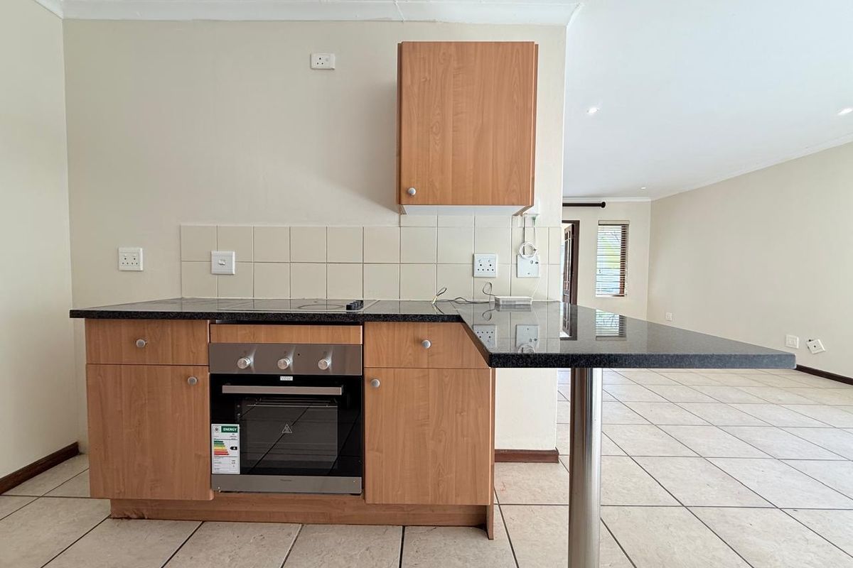 A compact kitchen area featuring laminate cabinetry, granite countertops with a breakfast bar, built-in oven, and tiled flooring, opening into an adjacent living space.