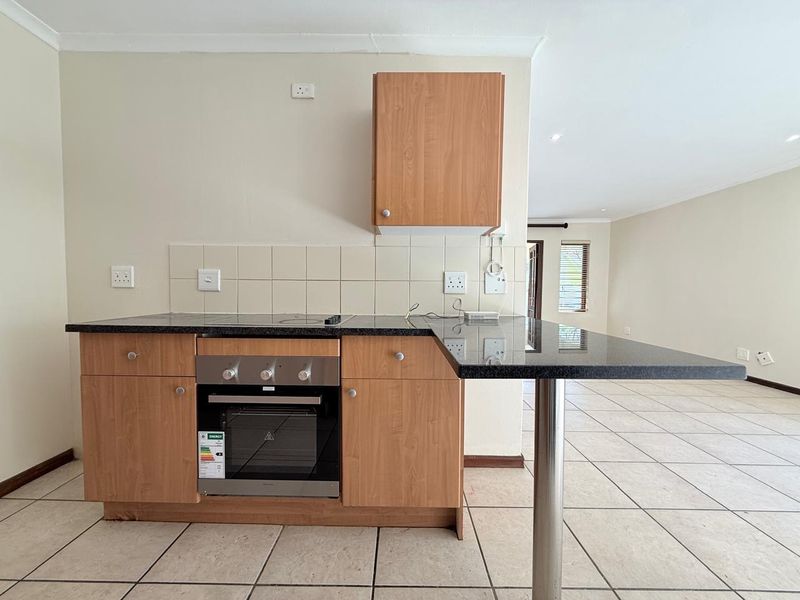 A compact kitchen area featuring laminate cabinetry, granite countertops with a breakfast bar, built-in oven, and tiled flooring, opening into an adjacent living space.