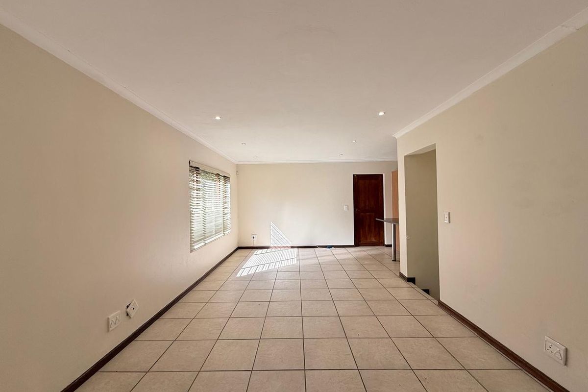 A spacious, empty living area featuring neutral walls, tiled flooring, recessed downlighting, and natural light through a window with fitted venetian blinds.