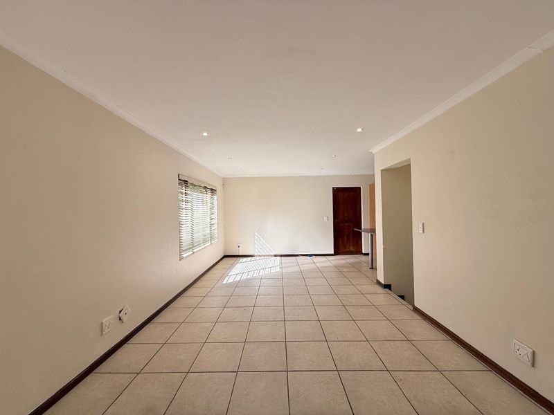 A spacious, empty living area featuring neutral walls, tiled flooring, recessed downlighting, and natural light through a window with fitted venetian blinds.