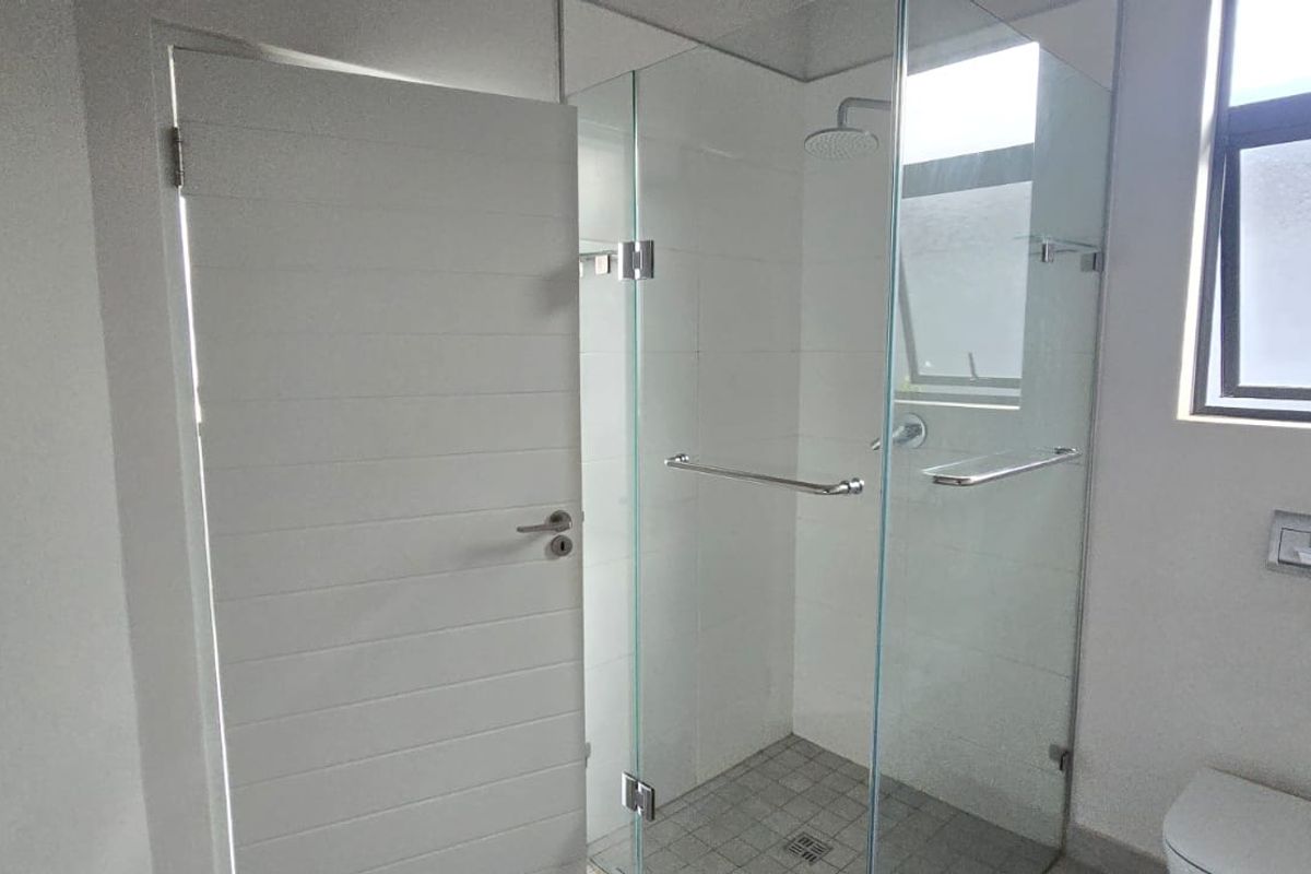 A clean, modern bathroom featuring a glass-enclosed shower with a rain shower head, wall-mounted toilet flush plate, and neutral tiling.
