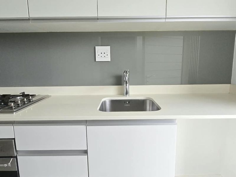 Modern kitchen area featuring white cabinetry, stone countertops, a gas hob, built-in oven, and a sleek glass splashback with a stainless steel sink.
