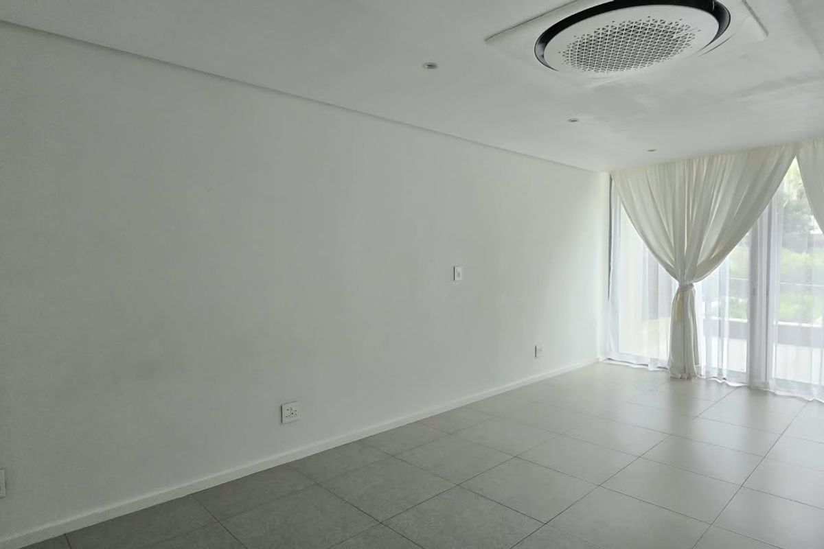 A bright, spacious, and empty living area featuring neutral tiled flooring, recessed lighting, a ceiling-mounted air conditioner, and large glass doors leading outdoors.