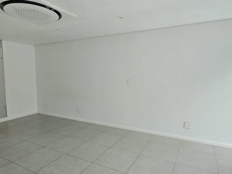 A bright, empty living area featuring neutral tiled flooring, recessed ceiling lighting, and a modern ceiling-mounted air conditioning unit.