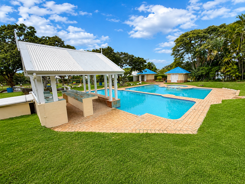 A well-maintained outdoor pool area featuring a covered entertainment pavilion with a built-in bar, surrounded by landscaped lawns and additional outbuildings.
