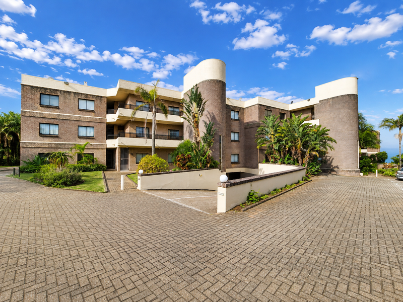 A well-maintained multi-story apartment complex featuring a brick facade, private balconies, and a paved driveway entrance with established landscaping.