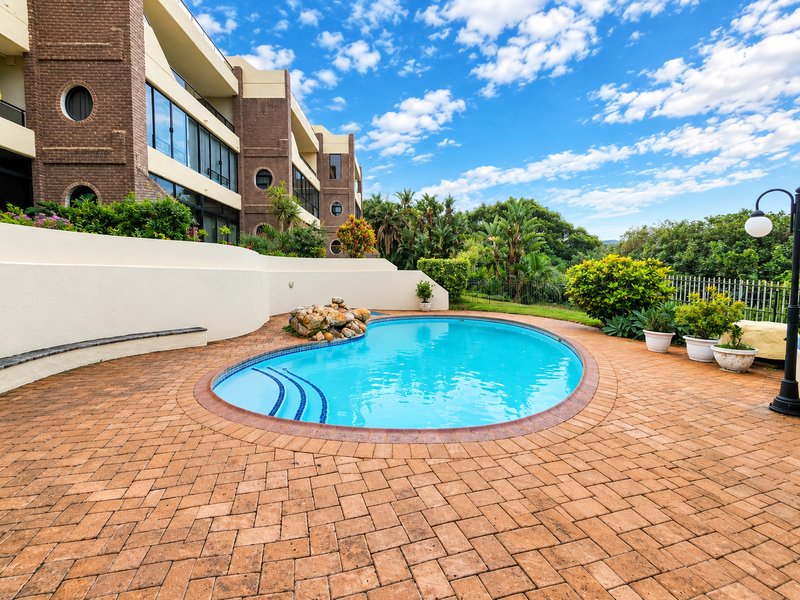 A well-maintained outdoor pool area featuring brick paving, a rock water feature, and a curved retaining wall set against a landscaped garden.