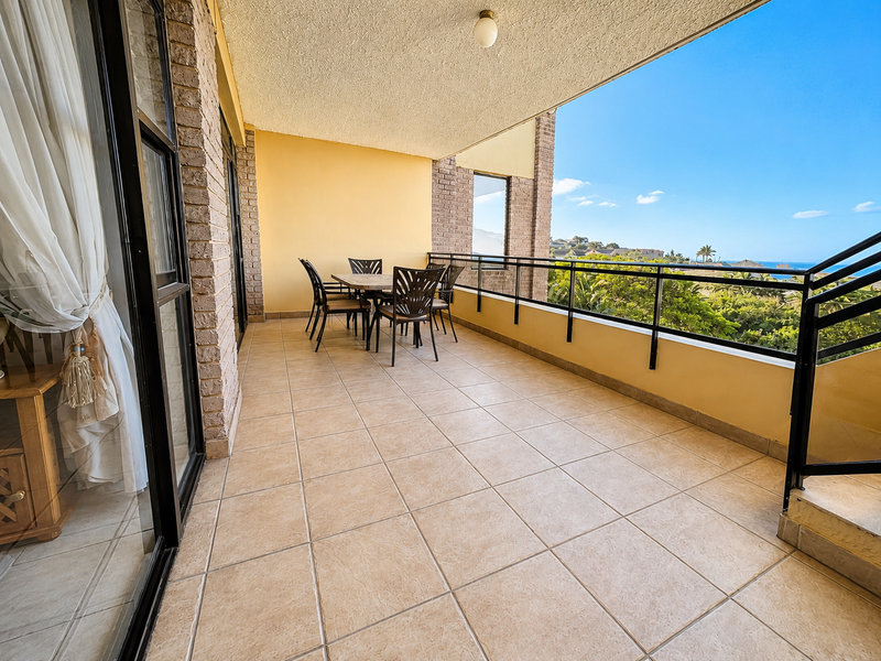 A spacious covered balcony featuring tiled flooring, brick accents, and a metal railing, offering an open outdoor area with scenic views.