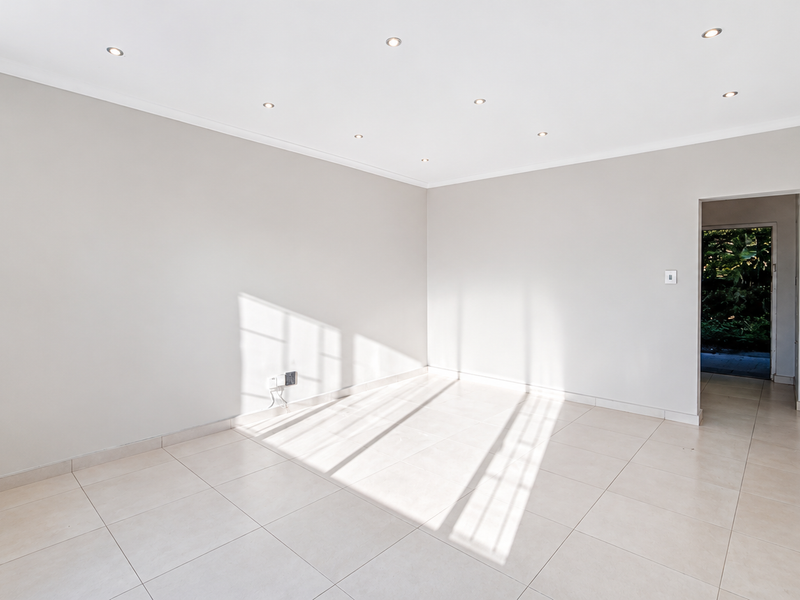 A bright, empty living area featuring neutral tiled flooring, recessed downlighting, and clean wall finishes, offering a versatile space for interior design.