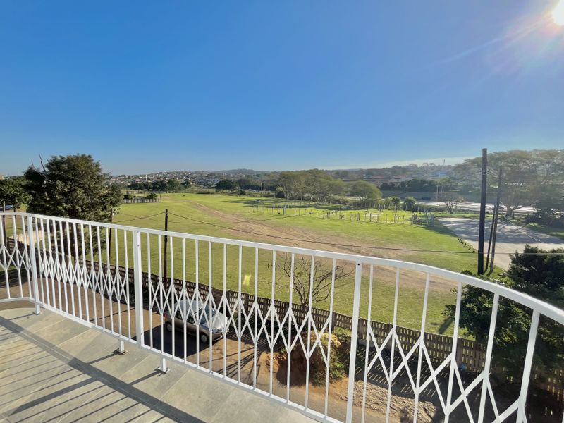 A spacious balcony featuring a white metal balustrade and tiled flooring, offering an expansive view over the surrounding neighborhood and green space.