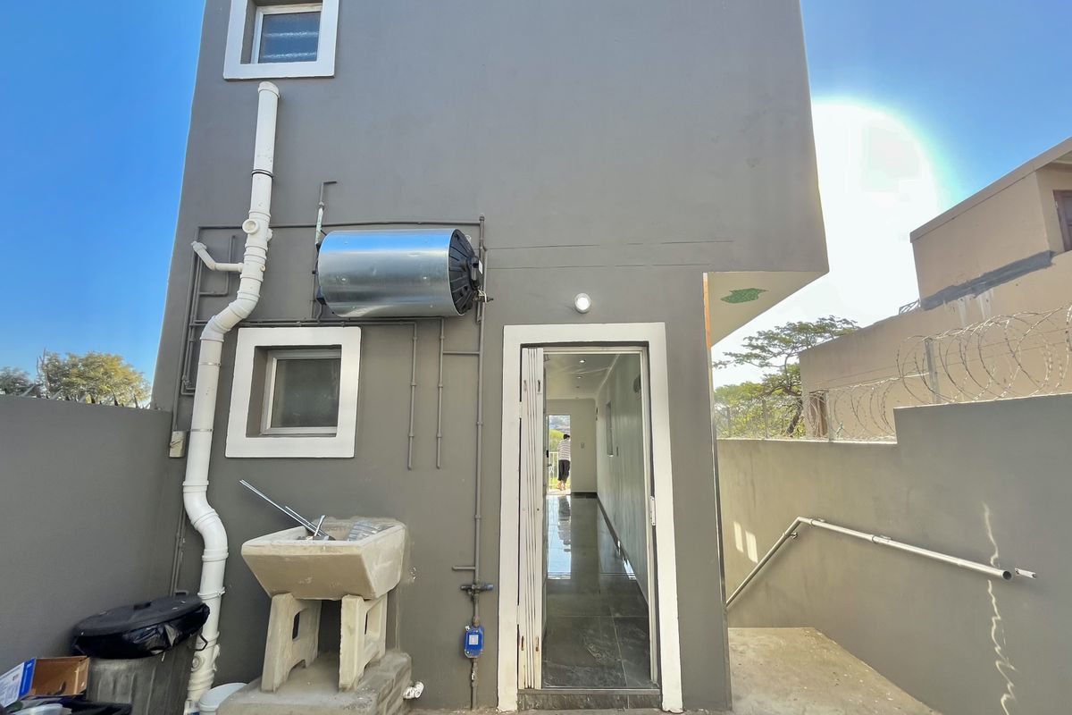 Rear exterior view of the apartment featuring an external wash trough, wall-mounted electric geyser, and a secure boundary wall with razor wire.