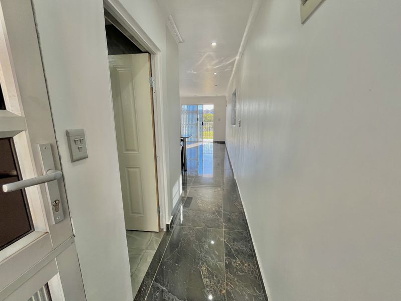 A bright, empty hallway featuring polished dark marble-look floor tiles, recessed downlighting, and a clear view through to the balcony.