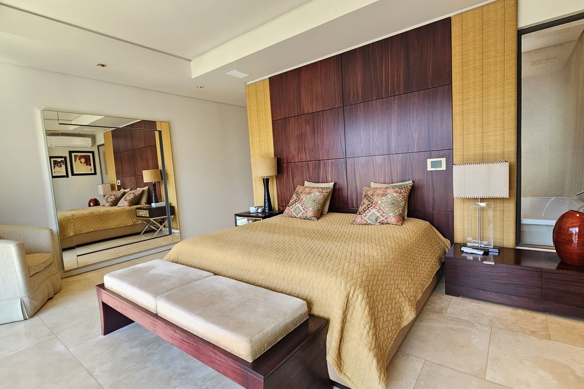Spacious bedroom featuring a custom wood-paneled headboard, stone tile flooring, recessed lighting, and an integrated glass partition leading to the en-suite bathroom.