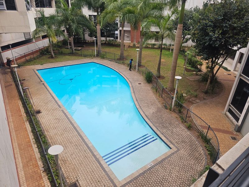 Elevated view of a communal swimming pool area featuring paved surrounds, landscaped gardens with palm trees, and secure perimeter fencing within an apartment complex.