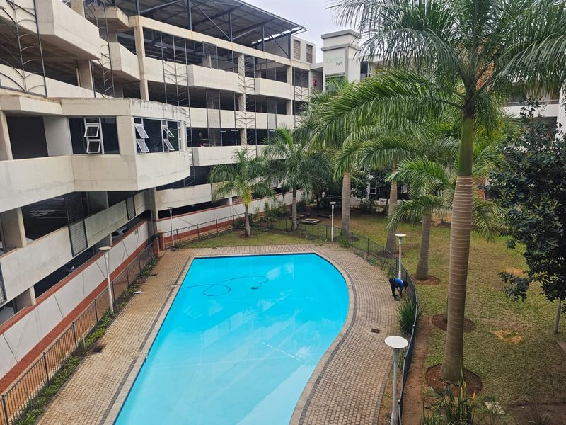 Communal swimming pool area featuring a paved surround, landscaped garden with palm trees, and adjacent multi-level parking structure.