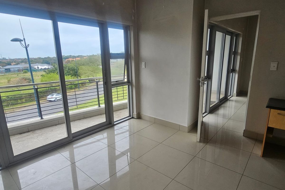An empty, light-filled living area featuring large sliding glass doors that open onto a balcony, with neutral tiled flooring throughout.