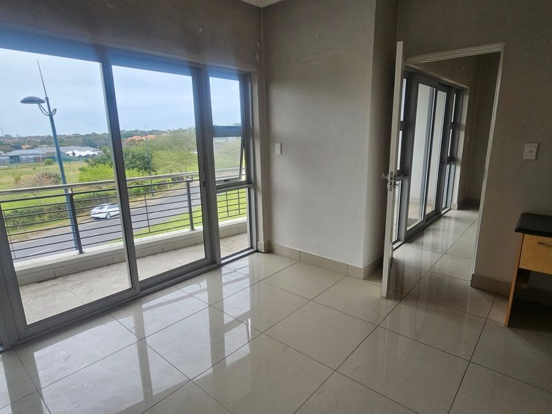 An empty, light-filled living area featuring large sliding glass doors that open onto a balcony, with neutral tiled flooring throughout.