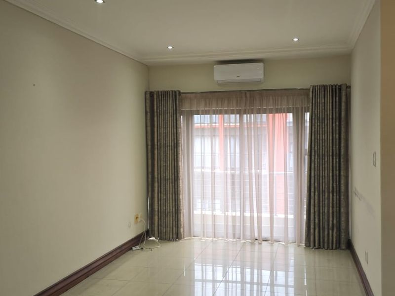 A bright, empty living room featuring tiled flooring, recessed downlights, an air conditioning unit, and a sliding glass door leading to a balcony.