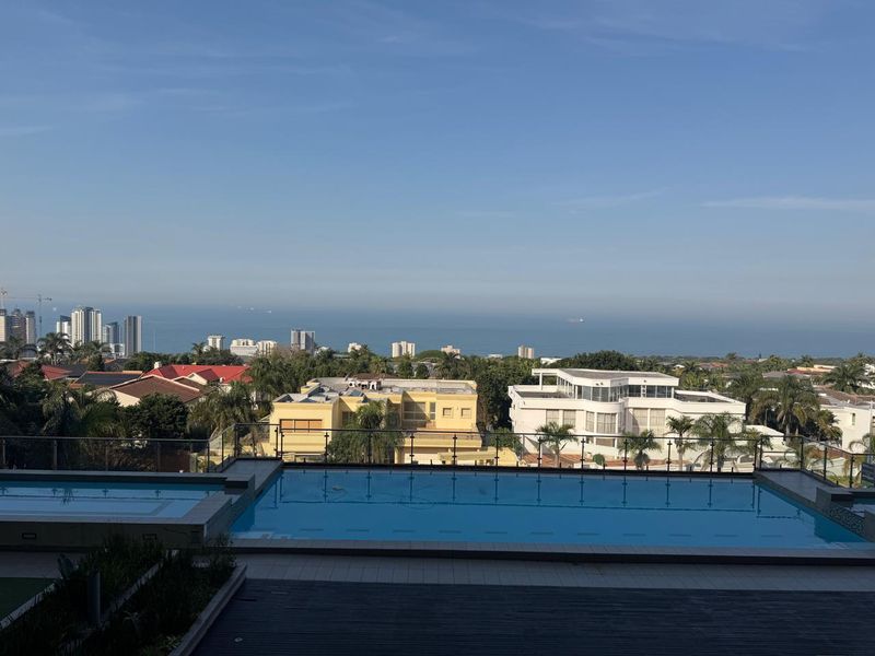 An elevated outdoor pool area featuring a glass balustrade and expansive coastal and city skyline views.
