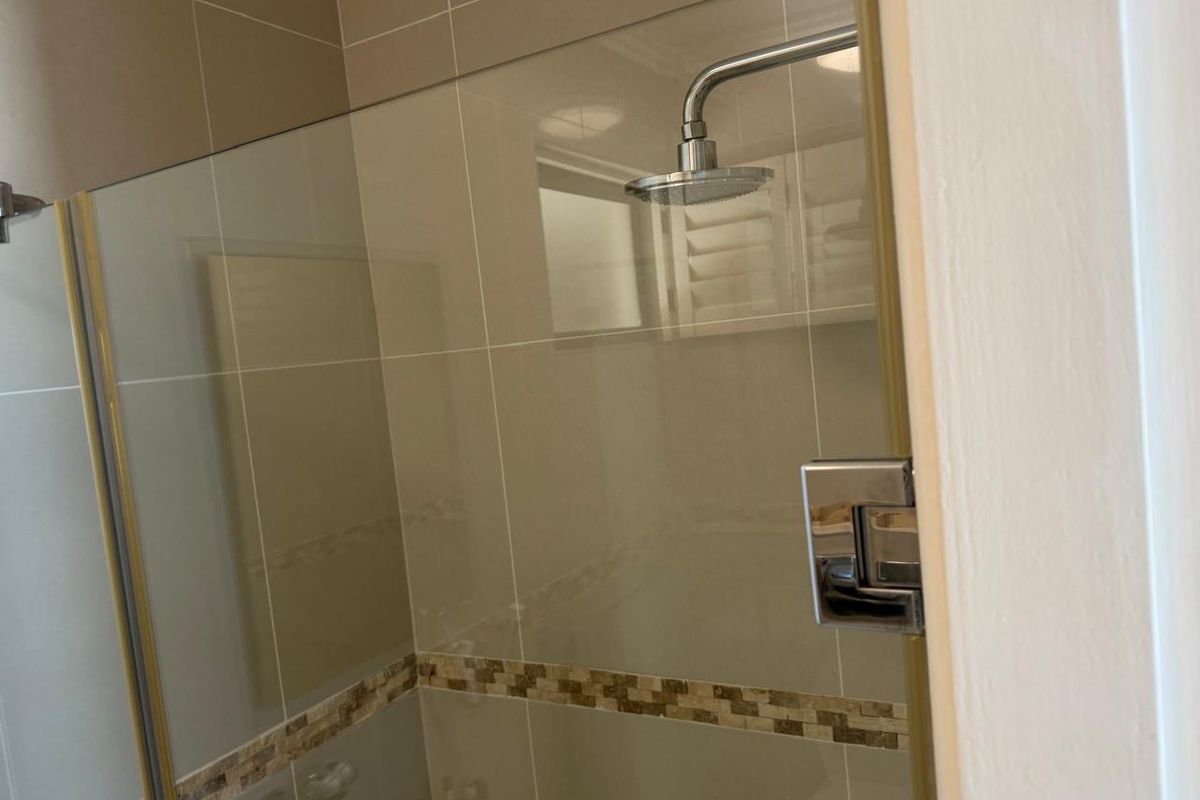A modern bathroom shower enclosure featuring a glass screen, rain shower head, and neutral-toned wall tiles with a decorative mosaic border.