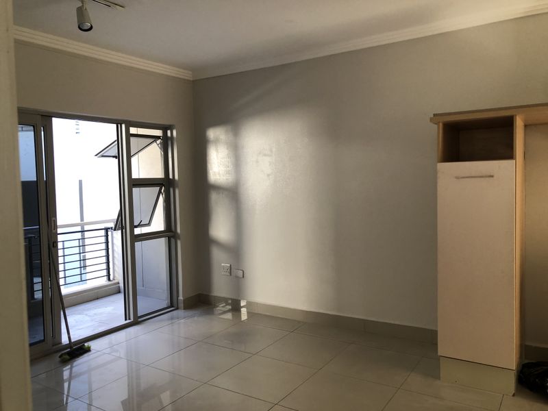 A bright, empty bedroom featuring tiled flooring, a built-in cupboard, and a sliding glass door providing access to a private balcony.