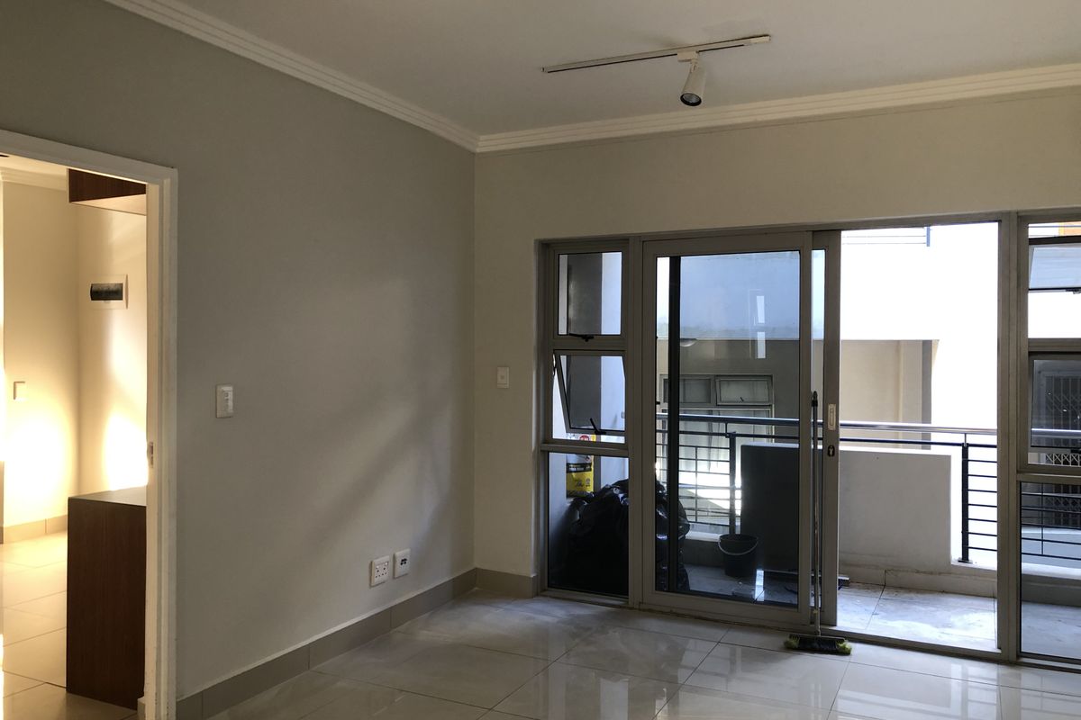 An empty, well-lit living area featuring neutral tiled flooring, modern track lighting, and a sliding glass door leading to an outdoor balcony.