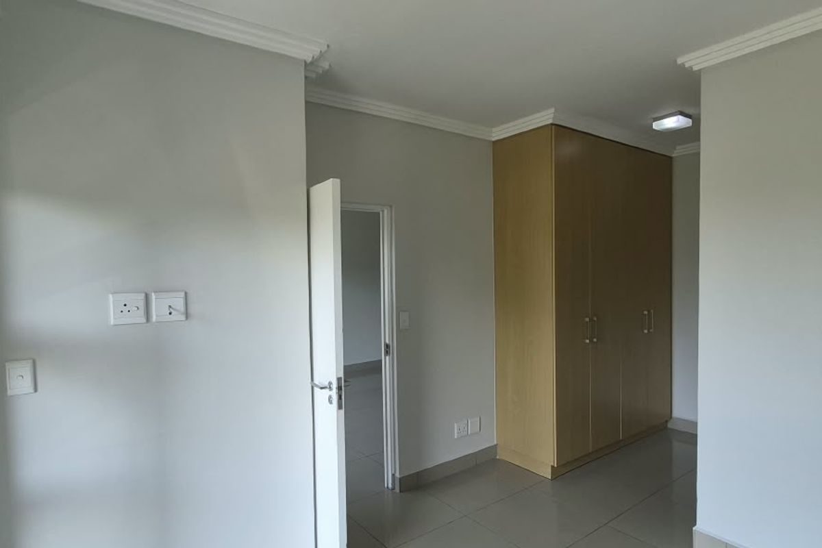 A bright, empty bedroom featuring neutral walls, tiled flooring, built-in wooden cupboards, and modern cornicing, offering a clean and versatile living space.
